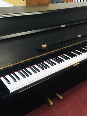 Manthey Klavier schwarz matt mit zwei Pedalen auf rotem Teppich, Frontansicht