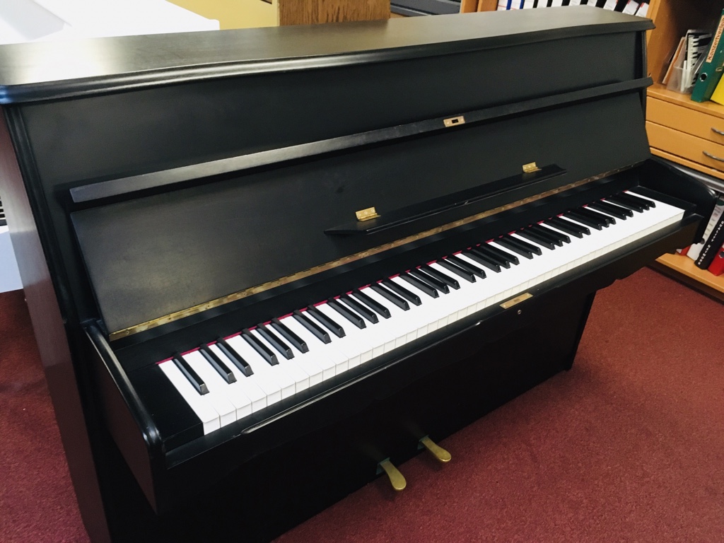 Manthey Klavier schwarz matt mit zwei Pedalen auf rotem Teppich, Frontansicht